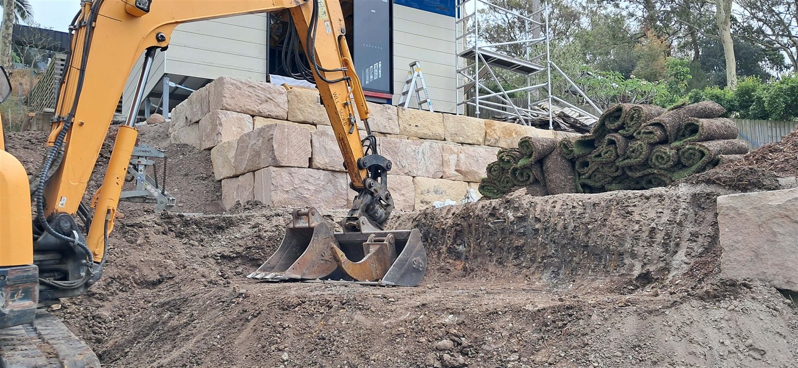 Excavator preparing sandstone retaining wall earthworks on a sloped residential site
